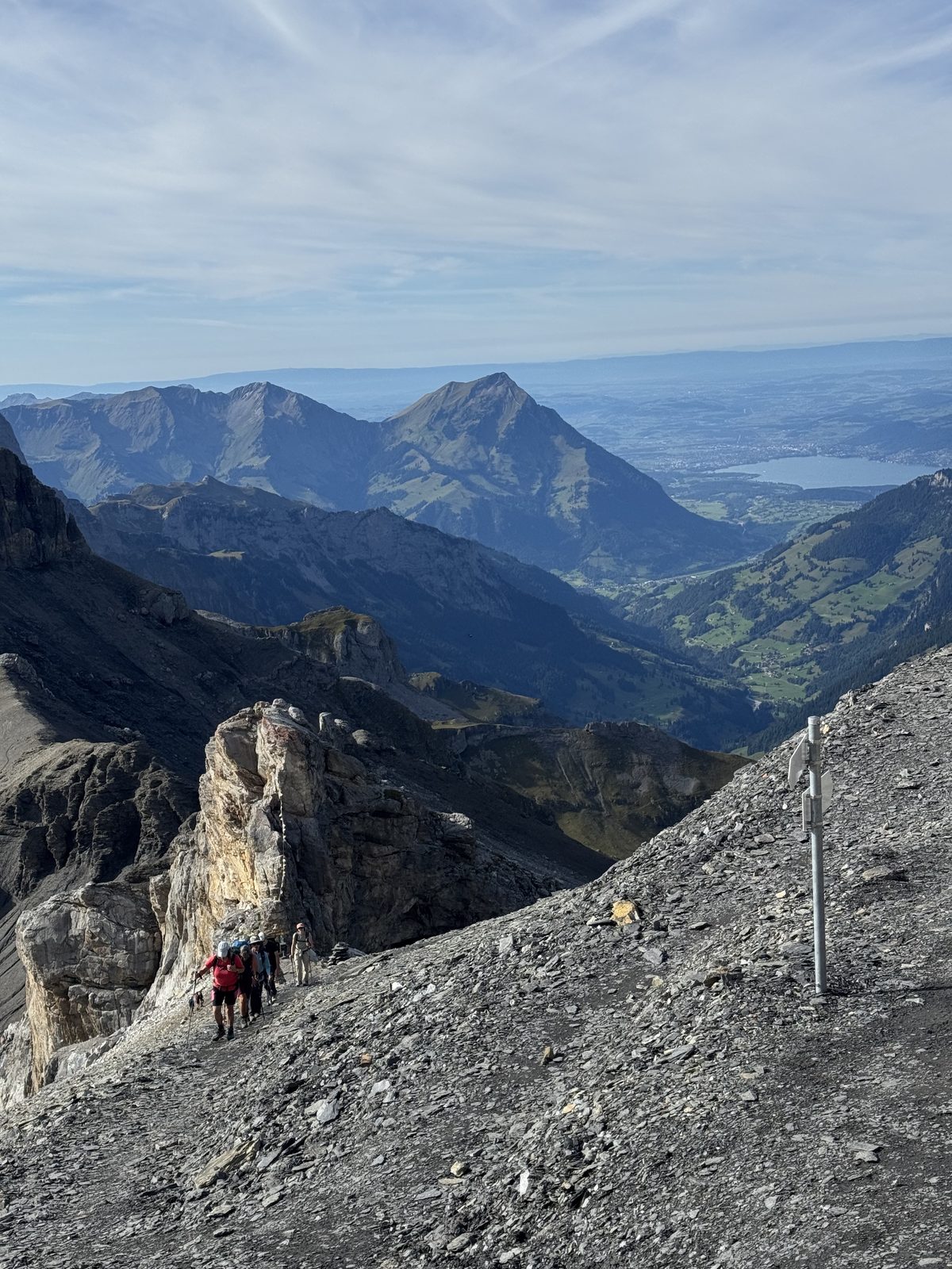 Group of hikers climbing the rocky trail towards the Hohtürli, with the Bernese valleys and a lake in the background